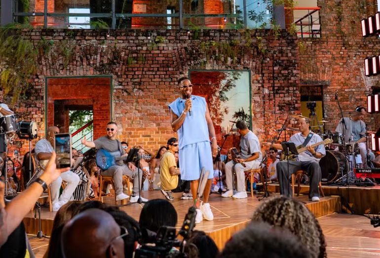 Mumuzinho cantando no palco do Parque Glória Maria, no Rio de Janeiro, durante o pôr do sol; ele sorri e interage com o público que está posicionado ao redor do palco em formato 360°.