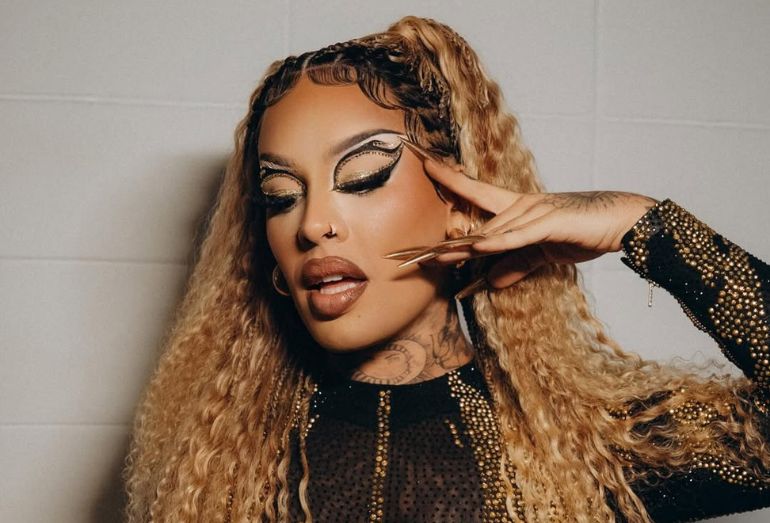 Fotografia em plano médio da artista Gloria Groove, uma drag queen de pele parda e longos cabelos loiros cacheados. Ela está de perfil parcial, com os olhos semicerrados, destacando uma maquiagem artística elaborada com delineado gráfico preto e dourado, além de pedrarias. Gloria veste um figurino preto de gola alta com transparências e aplicações de tachas douradas. Suas mãos estão próximas ao rosto, exibindo unhas postiças extremamente longas e douradas. O fundo é uma parede de azulejos brancos simples, criando um contraste com o visual luxuoso da artista.
