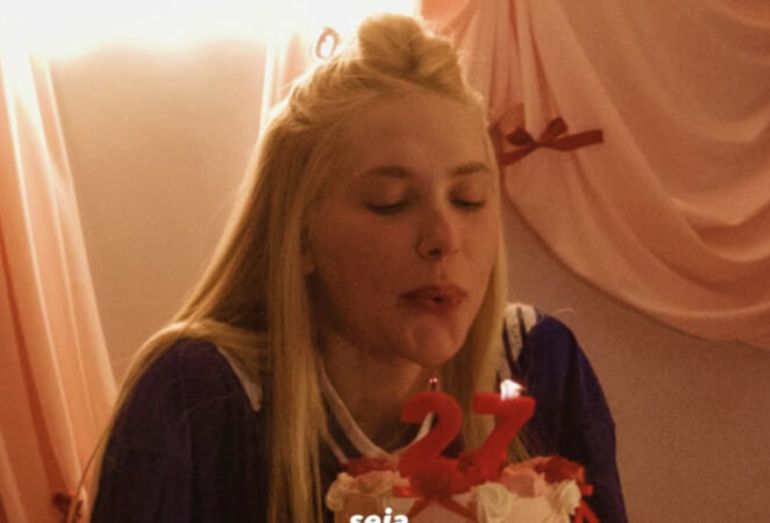 A influenciadora digital Virginia Fonseca, loira, agachada com os olhos semi-fechados e fazendo bico, soprando as velinhas acesas em um bolo de aniversário. O bolo tem as velas de número "27" em vermelho e é decorado com doces em tons de rosa. Virginia veste uma blusa roxa estilo jersey esportivo. O fundo é composto por cortinas em tons de rosa e detalhes em laços vermelhos.