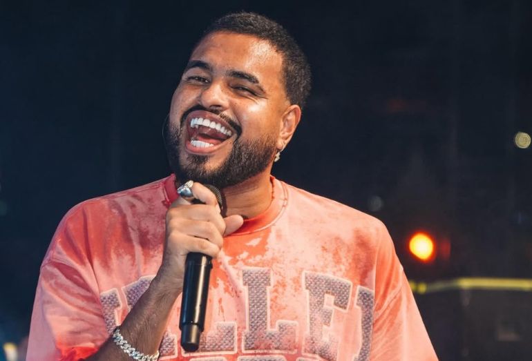 Fotografia em plano médio do cantor Yan, um homem negro de barba e cabelos curtos, em cima de um palco iluminado. Ele está rindo com a boca bem aberta, expressando grande felicidade, enquanto segura um microfone com a mão direita e canta. Veste uma camiseta tie-dye salmão e laranja, com letras grandes estampadas em relevo, e usa uma pulseira de elos prateada no pulso direito. O fundo é escuro, com pontos de luz e holofotes amarelos em desfoque.